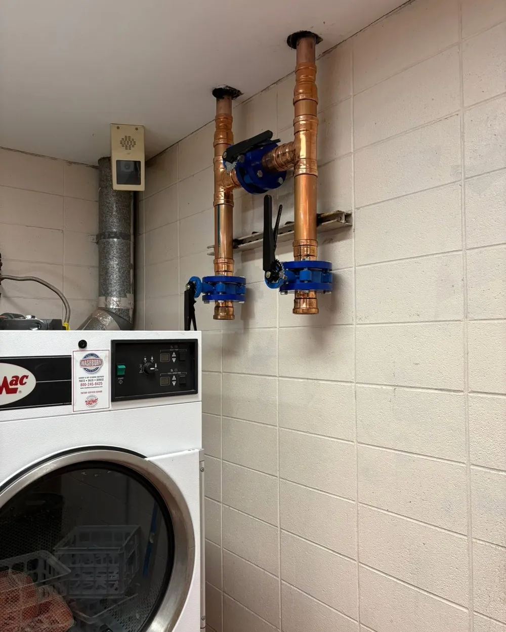 New copper piping with two blue butterfly valves installed on a block wall above commercial laundry machines